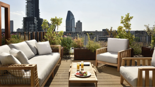 ミラノ ヴルティカレ ウナ エスペリエンツのルーフトップテラス。木製フレームの白いソファとチェアが並ぶモダンなパティオには、フルーツプレートとジュースが置かれ、背景にはミラノの象徴的な高層ビル群が広がっている