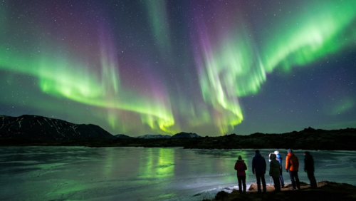 アイスランドの夜空に舞う緑と紫のオーロラ、前景に山と溶岩原、人々が冬服で鑑賞している様子
