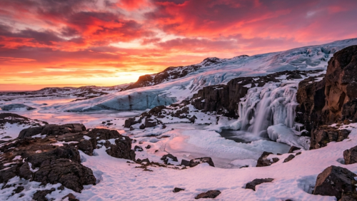 冬のアイスランド、赤く染まる夕焼け空の下に氷河と滝が広がる壮大な風景
