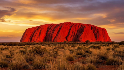 朝焼けで赤く輝くウルル（エアーズロック）と広大な砂漠、空は黄金色と紫色のグラデーション、オーストラリア中央部の大地の壮大な景色