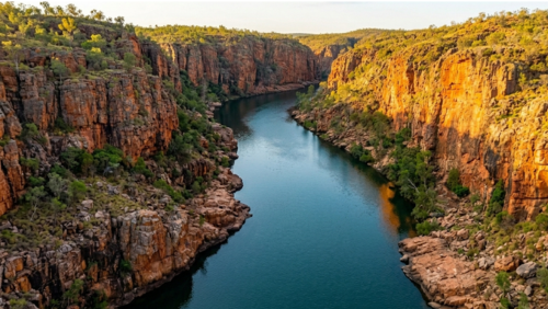 北オーストラリアのニトミルク国立公園にある、キャサリン渓谷（ニトミルク渓谷）の壮大な風景写真です。画面の両側には、約23億年前の地層が重なり合って形成された切り立った砂岩の断崖がそびえ立っています。岩肌は酸化した鉄分によって赤褐色やオレンジ色に輝き、夕日の光を受けて燃えるような質感をみせています。

断崖の間を縫うように、深く静かなキャサリン川がエメラルドグリーンから深い紺色のグラデーションを描きながら流れています。人工物は一切映っておらず、岩の裂け目に根を張る頑強な緑の木々だけが、この「地球の原風景」に彩りを添えています。太古から変わることのない、静寂と力強さに満ちた大自然のパノラマです。