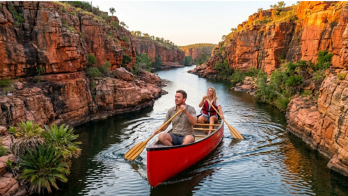 北オーストラリアのニトミルク国立公園にあるキャサリン渓谷で、赤いカヌーに乗って川を進む男女を近距離から捉えた臨場感あふれる写真です。

前景： 中央に大きく配置された赤いカヌーには、パドルを漕ぐ男性と女性が乗っています。水面にはカヌーが進むことで生じる穏やかな波紋が広がっています。

背景： 左右には、悠久の時を経て形成された赤い砂岩の巨大な断崖がそびえ立っています。岩肌には複雑な地層の筋が見え、夕日の光を受けてオレンジ色に輝いています。

雰囲気： 渓谷は奥の方まで曲がりくねりながら続いており、静寂の中に二人きりの「秘境感」が際立つ構成です。断崖の隙間には熱帯の植物やソテツのような緑が点在し、太古の地球の姿を彷彿とさせます。