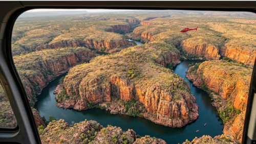 ヘリコプターの窓越しから見下ろした、ニトミルク国立公園（キャサリン渓谷）の壮大な空撮写真です。

前景： 画面の左右と上部には、ヘリコプターの機体内部と窓枠がわずかに映り込んでおり、実際に遊覧飛行を体験しているような視点を演出しています。

中景： 蛇行するキャサリン川が、赤茶色の巨大な砂岩の台地を深く切り裂きながら続いています。川は深いエメラルドグリーンで、切り立った断崖の形状に沿って「U字」や「S字」に大きくカーブしています。遠くの空には、別の赤いヘリコプターが小さく飛んでおり、景色のスケールの大きさを際立たせています。

背景： 地平線まで続く広大な低木林（ブッシュ）と、太古の地形が織りなす層状の岩壁が夕日に照らされ、オレンジ色から金色に輝いています。

雰囲気： 地上からは決して見ることができない、地球の脈動を感じさせるダイナミックな地形を一望できる、圧倒的な絶景です。