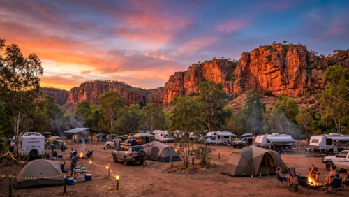 オーストラリア北部、ニトミルク国立公園（キャサリン渓谷）に実在する「ニトミルク・キャンプグラウンド」の敷地内から撮影した、臨場感あふれる夕暮れ時の風景写真です。

背景： 画面右側から奥にかけて、ニトミルク・ゴージを象徴する巨大な赤いサンドストーン（砂岩）の断崖がそびえ立っています。夕日の光を浴びた断崖は燃えるようなオレンジ色に輝き、その上の空はピンク、紫、オレンジが混ざり合う幻想的なサンセットのグラデーションに染まっています。

前景： 複数のグループがそれぞれのキャンプサイトで過ごしています。右下では焚き火を囲んで談笑する人々、左下ではテントの前で夕食の準備をする人物が写っています。キャンプサイトを繋ぐように飾られたストリングライト（電飾）やランタンが、温かい光を放ち始めています。

中景： 敷地内にはオフロード仕様の4WD車、キャンパーバン、大小様々なテントが並んでいます。地面はアウトバック特有の乾いた赤い砂土で、周囲にはユーカリやペーパーバークの木々が自生しており、自然と調和した宿泊施設の様子が描かれています。

雰囲気： 人工的な入り口を排除し、キャンプ場の内部から断崖を見上げる構図にすることで、大自然の懐に抱かれて夜を待つ、静かで贅沢な時間を表現しています。
