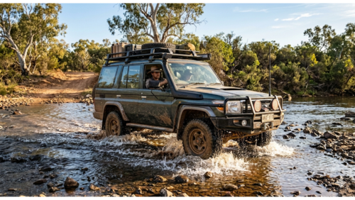 オーストラリアの未開の地（アウトバック）で、四輪駆動車が浅い川を横切る「ウォーター・クロッシング」の瞬間を捉えた、臨場感あふれる実写風の写真です。

車両の描写： 暗緑色のトヨタ・ランドクルーザー（70シリーズ）のオフロード仕様車が、画面中央で右から左へと川を渡っています。車体には、深い水深でも走行可能な吸気用のシュノーケル、頑丈なフロントバンパー（ブルバー）、大型のオフロードタイヤが装備されています。

装備と積載： ルーフキャリアには、予備の燃料缶（ジェリカン）や予備タイヤ、丸められたキャンプ用の寝具（スワッグ）などが積み込まれており、長距離の自給自足の旅を象徴しています。

動的な演出： タイヤが川底の石を噛み、周囲に白い水しぶきを豪快に上げています。泥の混じった水が車体に飛び散り、過酷な走行環境を物語っています。

背景と環境： 背景には、アウトバック特有のユーカリの木々が立ち並び、乾いた大地と川岸の岩が描かれています。空は明るく、柔らかな午後の光が車両と水しぶきを照らしています。

人物： 運転席には、オーストラリアのアウトバックらしい帽子を被った男性が座り、真剣な表情で前方の路面を注視しながらハンドルを握っています。