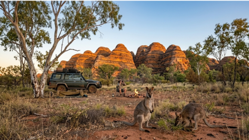 オーストラリアのパーヌルル国立公園（バングル・バングル）を背景に、野生のワラビーとキャンプシーンを捉えた実写風の写真です。

前景： 2匹の野生のワラビーが赤い砂土の上にいます。1匹は後ろ足で立ち上がり、好奇心旺盛な表情でこちらをじっと見つめており、もう1匹は隣で地面の草を食べています。ワラビーの柔らかな毛並みや長い尾が鮮明に描写されています。

中景： 画面左側には、オフロード仕様の暗緑色の4WD車が停車しており、その横には丸められた寝具（スワッグ）が置かれています。中央奥では、帽子を被った男性がキャンプチェアに座り、双眼鏡で静かにワラビーを観察しています。足元では小さな焚き火から煙が上がっています。

背景： 公園の象徴である、オレンジと黒の縞模様が特徴的な「蜂の巣ドーム（バングル・バングル山脈）」が画面中央に大きくそびえ立っています。周囲には白い幹のユーカリの木々が立ち並び、午後の柔らかな光が風景全体を温かく照らしています。