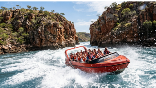 西オーストラリア州の秘境、ホリゾンタル・フォールズ（水平の滝）の激流を突き進む高速ジェットボートを捉えた、躍動感あふれる実写風の写真です。

中央の被写体： オレンジ色の船体が鮮やかな大型のジェットボートが、画面右下から中央へ向かって猛スピードで進んでいます。ボートのフロント部分には「HORIZONTAL FALLS ADVENTURES」という白いロゴが刻まれています。

乗客の様子： ボートにはオレンジ色のライフジャケットを着用した多くの観光客が乗っており、前方の激流を指さしたり、興奮した表情で歓声を上げたりしています。

激流の描写： ボートの周囲には、潮の満ち引きによって生じた激しい白波と渦が巻いています。背後の狭い岩の隙間からは、大量の海水が滝のように勢いよく流れ出しており、水しぶきが舞っています。

背景と環境： 左右には荒々しい赤褐色の砂岩の崖がそびえ立ち、崖の上にはまばらな緑の植生が見えます。空は澄み渡った青空で、太陽の光が水面と白いしぶきをキラキラと輝かせています。

アングル： 低い位置からのローアングルで撮影されており、迫りくる水のパワーとボートのスピード感が強調されています。