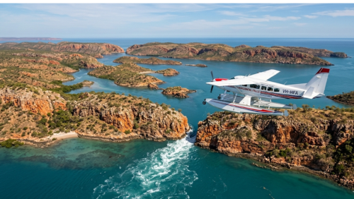 西オーストラリア州の秘境、ホリゾンタル・フォールズ（水平の滝）の上空約1000フィートから捉えた、壮大なパノラマ空撮写真です。

前景の飛行機： 画面中央右側には、白い機体に赤と青のライン、そして「HORIZONTAL FALLS ADVENTURES」と登録記号「VH-HFA」の文字がはっきりと刻まれた、セスナ 208 キャラバン アンフィビアン（水陸両用シープレーン）の全体像が、翼を大きく広げて飛行しています。窓越しにパイロットと数人の乗客（オレンジ色のライフジャケット着用）の姿が見えます。

背景の地形： 下方には、澄んだターコイズブルーの海水が、数百もの無人島、入り組んだ入り江、そして切り立った赤い砂岩の崖を囲む、非常に複雑な海岸線が広がっています。島々には乾燥した植生が点在しています。

見どころ（滝）： 飛行機の直下には、ホリゾンタル・フォールズの象徴である、2つの巨大な崖の隙間を強力な潮の満ち引きによる海水が「水平の滝」となって白い激流と渦を作りながら突き抜けていく様子が、はっきりと描写されています。

遠景と雰囲気： 遠くには広大なインド洋の水平線が澄み渡った青空の下に広がり、島々の影が水面に落とされています。岩の質感、水面のきらめき、飛行機の細部まで鮮明に表現した、臨場感あふれる超高解像度8K画像です。