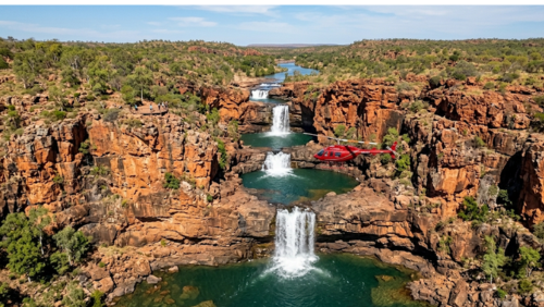 西オーストラリア州のキンバリー地域北部にある、ミッチェル・フォールズの4段に重なる滝を上空から捉えた、ダイナミックな空撮写真です。

滝の構造と色彩： 荒々しい赤褐色の砂岩が階段状に削られ、4つの独立した滝が垂直に流れ落ちています。それぞれの滝の麓には、透き通ったエメラルドグリーンの深い滝壺（プランジ・プール）が形成されており、白い水しぶきと美しい対比を生んでいます。

ヘリコプターの存在： 画面中央の滝のすぐ近くを、赤い機体のヘリコプターが飛行しており、この滝の巨大なスケール感を強調しています。

周囲の環境と人物： 峡谷の周囲には乾燥した低木やユーカリの樹木が点在しています。画面左側の崖の上にある木製の展望デッキには、数人の観光客の姿が小さく見え、この大自然の絶景を見下ろしています。

遠景： 背景にはキンバリー地域特有の広大な赤土の平原と丘陵地帯が、澄み切った青空の下、地平線の彼方まで続いています。

全体の雰囲気： 人里離れた「陸の孤島」としての野生味と、自然が造り出した幾何学的な美しさが共存する、超高解像度8K画像です。