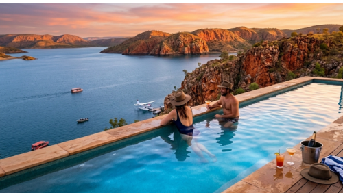 西オーストラリア州のレイク・アーガイル・リゾートにある、世界的に有名なインフィニティ・プールから眺める夕暮れの風景写真です。

中央の被写体： プールの水面が、眼下に広がる巨大なアーガイル湖と地平線で溶け合うように繋がって見えます。プールの中では、アクーブラハットを被った女性と、カクテルを手にした男性のカップルが、縁に寄りかかってリラックスしながら絶景を眺めています。

背景の景観： 湖の周囲には、夕日に照らされて黄金色やオレンジ色に輝く荒々しい赤褐色の岩山（砂岩の崖）が連なっています。広大な湖面には島々が点在し、遠くには観光ボートや停泊中の水上飛行機（シープレーン）が小さく見えます。

プールサイドの演出： 手前の木製デッキには、トロピカルカクテルやワインクーラーに入ったシャンパン、ストライプのビーチタオル、そして予備のアクーブラハットが置かれ、贅沢なリゾートの時間を演出しています。

全体の雰囲気： 空は淡いピンクとオレンジ色の夕焼けに染まり、水面にはその色が美しく反射しています。ワイルドな大自然と、洗練されたリゾート体験が融合した、静寂と幸福感に満ちた超高解像度画像です