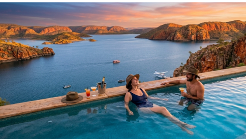 アーガイル湖のインフィニティ・プールの別バージョン、さらに人物の表情やリラックスした空気感が伝わる画像の代替テキストです。

代替テキスト（アクセシビリティ用）
西オーストラリア州の「ディスカバリーリゾーツ - レイクアーガイル」にある、伝説的なインフィニティ・プールを別角度から捉えた幻想的な風景写真です。

中央の被写体（カップル）： プールの水面に肩まで浸かり、カクテルを楽しみながらリラックスしている男女のカップルが描かれています。女性はアクーブラハットを被り、空を見上げて微笑んでおり、男性もまた満足げな表情で彼女に向き合っています。

背景の広がり： プールの縁から先には、穏やかなアーガイル湖の青い水面が地平線まで広がっています。湖に浮かぶ島々や、対岸の荒々しくも美しい赤い砂岩の崖が、沈みゆく夕日の光で黄金色に輝いています。

水面のディテール： 夕焼け空のオレンジやピンク、紫色のグラデーションが湖面とプールの水面に反射し、鏡のような美しさを作り出しています。遠くにはサンセットクルーズのボートや水上飛行機が小さく見え、この地が特別な休暇の場所であることを示唆しています。

プールサイドの小物： プールの縁には、トロピカルなカクテルグラス、冷えたシャンパンボトル、そしてもう一つのアクーブラハットが置かれ、贅沢で穏やかな時間の流れを象徴しています。

全体の雰囲気： 「いつか絶対に訪れたい」と思わせるような、大自然の雄大さとリゾートの快適さが完璧に調和した、超高解像度8K画像です。