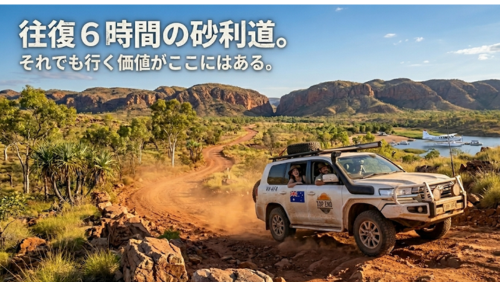 オーストラリアのアウトバック、乾燥した赤土の未舗装路（砂利道）を走行する、頑丈な白いトヨタ・ランドクルーザー200（4WD）のダイナミックな写真です。車は巻き上がる砂塵を切り裂きながら、画面中央を力強く進んでいます。車内には、アクーブラハットを被った男性と女性が笑顔で座っており、冒険を楽しんでいる様子です。車体は砂埃で汚れ、シュノーケル、ルーフラック、オフロードタイヤ、LEDライトバー、そしてオーストラリア国旗のステッカーや「TOP END」のロゴが確認できます。背景には、乾燥したブッシュ（スピニフェックスグラス、ユーカリの木）、そして遠くに広大な赤褐色の山々と、湖に浮かぶ水上飛行機（セスナ機）が見え、壮大なスケール感を感じさせます。画像の左上には、白い日本語の文字で「往復6時間の砂利道。それでも行く価値がここにはある。」と記されています。自然光、超高解像度画像です。