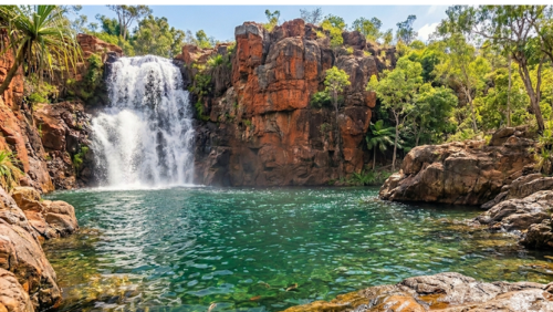 リッチフィールド国立公園にある「ワンギ・フォールズ」の壮大な自然景観です。画面左側では、高く切り立った赤褐色の砂岩の絶壁から、勢いよく白いしぶきを上げる滝がエメラルドグリーンの広大な天然プールへと流れ落ちています。周囲はパンダナスやシダ、ユーカリといった青々と茂る熱帯雨林に囲まれており、前景には濡れた質感の岩場が広がっています。人工物のない、手つかずの野生の美しさと静寂を感じさせる超高解像度8K画像です。