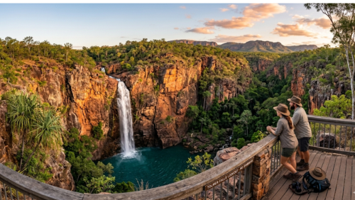リッチフィールド国立公園の「トルマー・フォールズ」を展望台から見渡す、ドラマチックな風景写真です。画面右側の木製展望台では、アクーブラハットを被ったカップルが手すりに寄りかかり、深い峡谷の向こう側に流れる滝を静かに見守っています。切り立った赤褐色の絶壁から一条の白い滝が、深いエメラルドグリーンの滝つぼへと垂直に流れ落ちており、周囲は鬱蒼とした熱帯雨林の緑に包まれています。手すりには「TOLMER FALLS LOOKOUT - NO SWIMMING」と刻まれており、夕暮れ時の黄金色の光が絶壁と山脈を赤く染め上げている、静寂と畏敬の念を感じさせる超高解像度8K画像です。