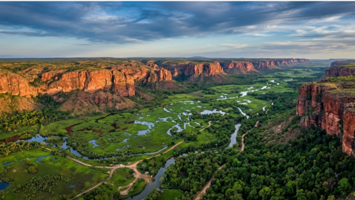 オーストラリア北部・カカドゥ国立公園を彷彿とさせる、手つかずの大自然の空撮写真。夕暮れ時の柔らかな光に照らされた赤い断崖絶壁が地平線まで続き、その下にはエメラルドグリーンの湿地帯と生命力あふれる熱帯雨林、ゆったりと流れる川が広がる神秘的な風景