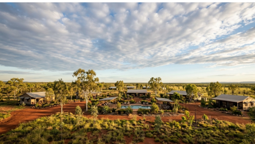 オーストラリア北部ノーザンテリトリー、バチェラーに位置する「リッチフィールド・アウトバック・リゾート」の広大な敷地。赤土の大地とユーカリの木々に囲まれ、ゆったりと配置されたコテージ風の建物、中央のプールエリア、そして地平線まで続くドラマチックな青空が広がる、開放感あふれるアウトバックのリゾート風景