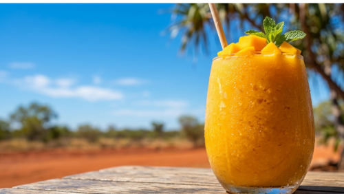 オーストラリア北部の鮮やかな青空と赤土の大地を背景に、木製のテーブルに置かれた濃厚なマンゴー・スムージー。結露した透明なグラスに注がれた鮮やかなオレンジ色のスムージーには、完熟マンゴーのカットとミントが添えられ、暑い日差しの中で喉を潤す最高の一杯を表現した料理写真