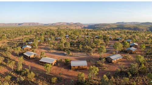 ニトミルク国立公園の広大なアウトバックに点在する、木製キャビンの宿泊施設を上空から捉えたドローン写真。赤土の大地とサバンナの低木に囲まれ、ゆったりと間隔をあけて配置されたキャビンが、朝の柔らかな光の中で自然と調和している様子を描いた全景ショット
