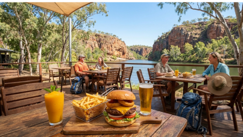 オーストラリア・ニトミルク国立公園のビジターセンターにある、川を一望するテラス席でのランチ。手前の木製テーブルには、分厚いハンバーガーと山盛りのポテト、マンゴースムージー、冷えたビールが並び、背景には赤い砂岩の断崖とユーカリの森が広がる。カジュアルな装いの旅行者たちがくつろぐ、活気と開放感にあふれたカフェ風景