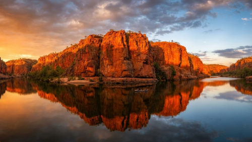 夕陽を浴びて燃えるようなオレンジ色に輝く、オーストラリア・ニトミルク渓谷の巨大な赤い砂岩の断崖。蛇行する静かな川面には、ドラマチックな空の雲と断崖のシルエットが鏡のように美しく反射している。広大な峡谷の中に小さなカヌーがポツンと浮かび、太古から続く自然の圧倒的なスケールと静寂を捉えたパノラマ写真