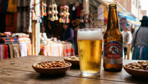 ボリビアの地元バーや屋台の木製テーブルに、泡立つボリビア産ビールのグラスと瓶、横にスパイシーに味付けされたナッツや豆類の小皿が並ぶグルメセット。背景には屋台のカラフルな装飾や地元の人々がぼんやり映り、自然光で色彩と質感が鮮明に映えるフォトリアル写真。