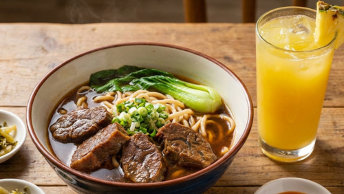 深めの丼に入った台湾の牛肉麺。柔らかく煮込んだ牛肉とコシのある麺、香味豊かなスープが見える。横には透明なグラスに注いだフレッシュなパイナップルジュースが置かれており、熱いスープと冷たいジュースのコントラストが楽しめる横長の画像。