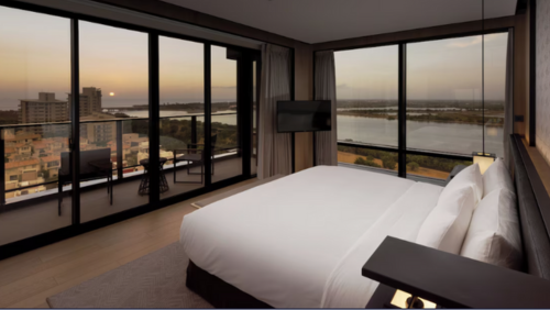 クラウンプラザ台南のジュニアスイート。高級寝具と羽毛の掛け布団を備えたキングサイズベッドが配置され、角部屋ならではの大きな二面採光の窓からは塩水渓のリバービューが一望できる。夕日が沈む川の景色を眺められるバルコニーと、室内にはセーフティボックスやテレビを完備した、ゆとりあるモダンな空間