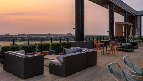 アロフト タイナン アンピンの屋上テラス。夕焼けに染まる空と台南の街並みを一望できる開放的な空間に、ゆったりとしたラタン調のソファセットやオレンジのハイチェアが配置されている。心地よい風を感じながらドリンクを楽しめる、モダンでスタイリッシュなルーフトップバー