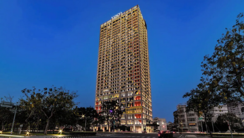 夕闇に包まれた嘉義の街にそびえ立つ「voco 嘉義（嘉義福容 voco 酒店）」の高層ビル外観。建物の窓から漏れる温かな光と、最上部の「voco」のロゴがライトアップされている。近代的な建築デザインが、深い青色の空を背景に美しく浮かび上がる夜景シーン