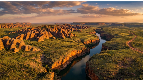 夕陽に照らされた西オーストラリア・キンバリー地方の広大な秘境風景。蛇行する深い峡谷と川、特徴的な縞模様のバングル・バングルの奇岩群、そして地平線へと続く赤土の未舗装路を一望するドローン空撮。ドラマチックな黄金色の光が大地を包み、太古から続く圧倒的な大自然のスケールを捉えたシネマティックな風景写真