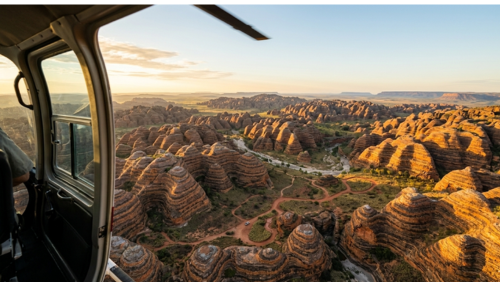 ヘリコプターの機内から見下ろした、パーヌルル国立公園の壮大なバングル・バングル山脈。開いたドア越しに、地平線まで続く縞模様の奇岩ドーム群と、赤土の道を走る小さな四輪駆動車が見える。空撮ならではの視点で、異世界のような広大な地形と冒険のスケールを捉えたドラマチックな写真