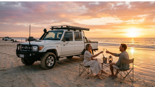 西オーストラリア州ブルームのケーブルビーチで、白い四輪駆動車（4WD）を砂浜に止め、夕日を眺めながらシャンパンを楽しむ男女。黄金色のサンセットがインド洋を照らし、波打ち際に置かれた小さなテーブルと椅子が「ワイルドで贅沢な」旅のひとときを演出している。背景には、砂浜を走る他の4WD車と、淡いピンクとオレンジに染まる夕焼け空が広がる風景写真