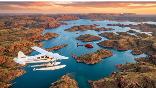 西オーストラリア州・アーガイル湖を上空から捉えた空撮写真。複雑に入り組んだ赤い大地と、無数に浮かぶ島々が「水の迷宮」のような景観を作り出している。手前には白いシープレーン（水上飛行機）、中央には赤いヘリコプターが飛行しており、広大な湖の上を遊覧している様子がわかる。夕陽が大地を赤く照らし、青い湖水との対比が美しい、圧倒的なスケール感の絶景写真。