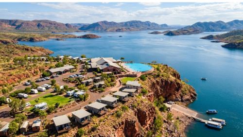 西オーストラリア州アーガイル湖の断崖絶壁に立つ「ディスカバリーリゾーツ・レイクアーガイル」の全景。赤い岩肌の岬に、象徴的なインフィニティ・プール、キャビン型の宿泊棟、キャンピングカーサイトが機能的に配置されている。背景には、真っ青な湖水に無数の島々が浮かぶ「水の迷宮」のような絶景が地平線まで広がり、湖畔にはボートの発着場も見える、圧倒的な開放感の空撮写真。