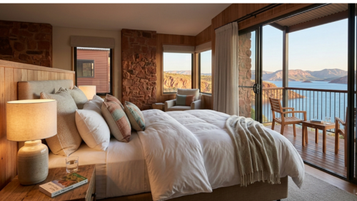 ディスカバリーリゾーツ・レイクアーガイルの贅沢なゲストルームの室内。清潔で柔らかな白いリネンが整えられた大きなベッドがあり、傍らには温かな光を灯すベッドサイドランプが置かれている。大きく開かれたスライド式のガラス扉の向こうには専用のプライベートバルコニーが広がり、真っ青なアーガイル湖と険しい赤い岩山の絶景を、ベッドに寝転んだまま眺めることができる開放的な空間。