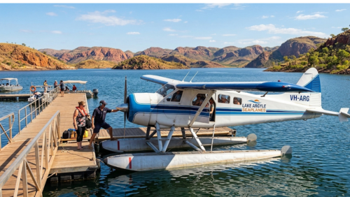 アーガイル湖の桟橋に係留された、「LAKE ARGYLE SEAPLANES」のロゴが入った白と青の水上飛行機（シープレーン）。フロートを備えた機体の側では、パイロットと乗客が搭乗の準備をしており、背後には赤い岩肌の山々と広大な湖が広がっている。晴天の下、穏やかな湖面から空の旅へと出発する直前の様子を捉えた、臨場感のある写真