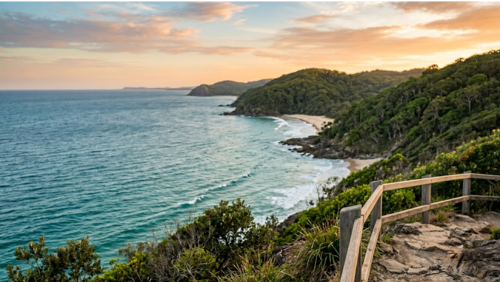 オーストラリア、ヌーサの海岸線を高台から望む風景。ターコイズブルーの海と白い波打ち際、右側にはラミントン国立公園を彷彿とさせる深い緑の熱帯雨林が広がっている。夕暮れ時の柔らかなピンクとオレンジの空が水面に映り、静寂で美しい自然の情景。