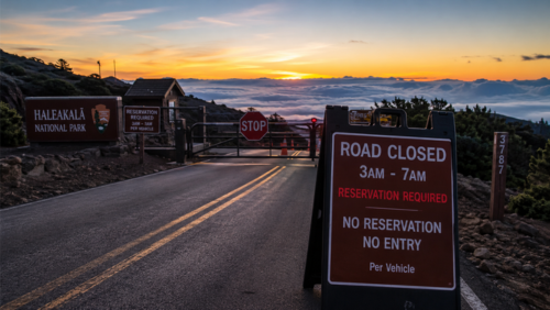 ハレアカラ国立公園の山道入口にあるゲートが閉鎖され、「ROAD CLOSED 3AM–7AM / RESERVATION REQUIRED / NO ENTRY」と書かれた看板が立っている早朝の風景。雲海の上に朝焼けの空が広がり、予約なしでは入園できない規制の厳しさを示している