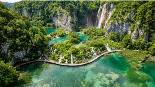 クロアチアのプリトヴィツェ湖群国立公園・下湖群の風景。エメラルドグリーンに輝く透明度の高い湖の上を木製の遊歩道が伸び、水底や泳ぐ魚が見えるほど澄んだ水が広がる。周囲は緑豊かな森と石灰岩の断崖に囲まれ、奥には大きな滝が流れ落ちている。