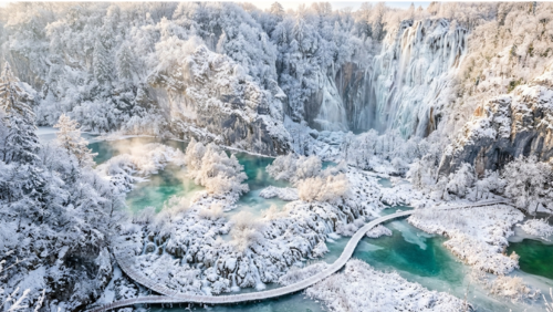 冬のプリトヴィツェ湖群国立公園。雪に覆われた森と湖の中で、滝が氷柱や氷のカーテンのように凍りついている。白銀の世界に包まれた遊歩道や木々の間から、青緑色の湖面がのぞき、静寂に満ちた幻想的な冬景色が広がっている