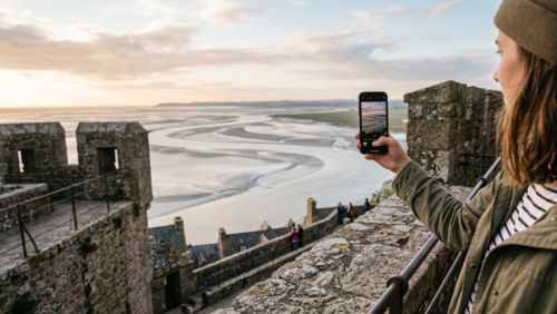 モン・サン・ミッシェルの城壁から湾と干潟の絶景をスマホで撮影する旅行者。島内ならではの見下ろし風景を楽しむ撮影スポットの様子